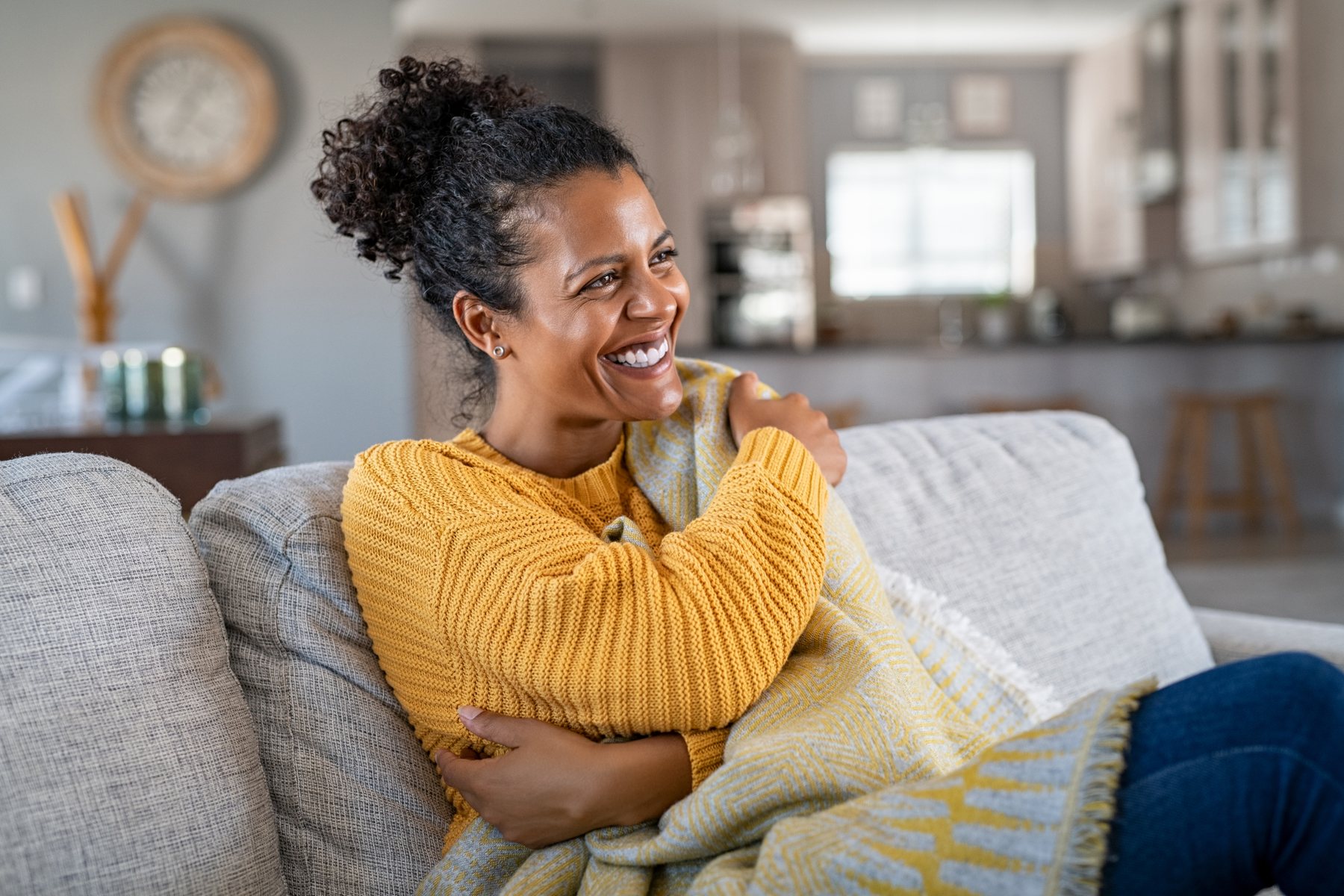 Beautiful black woman sitting on couch wrapped under blanket and laughing. Cheerful african american woman relaxing at home. Carefree and happy mid lady hugging herself with warm blanket in winter.