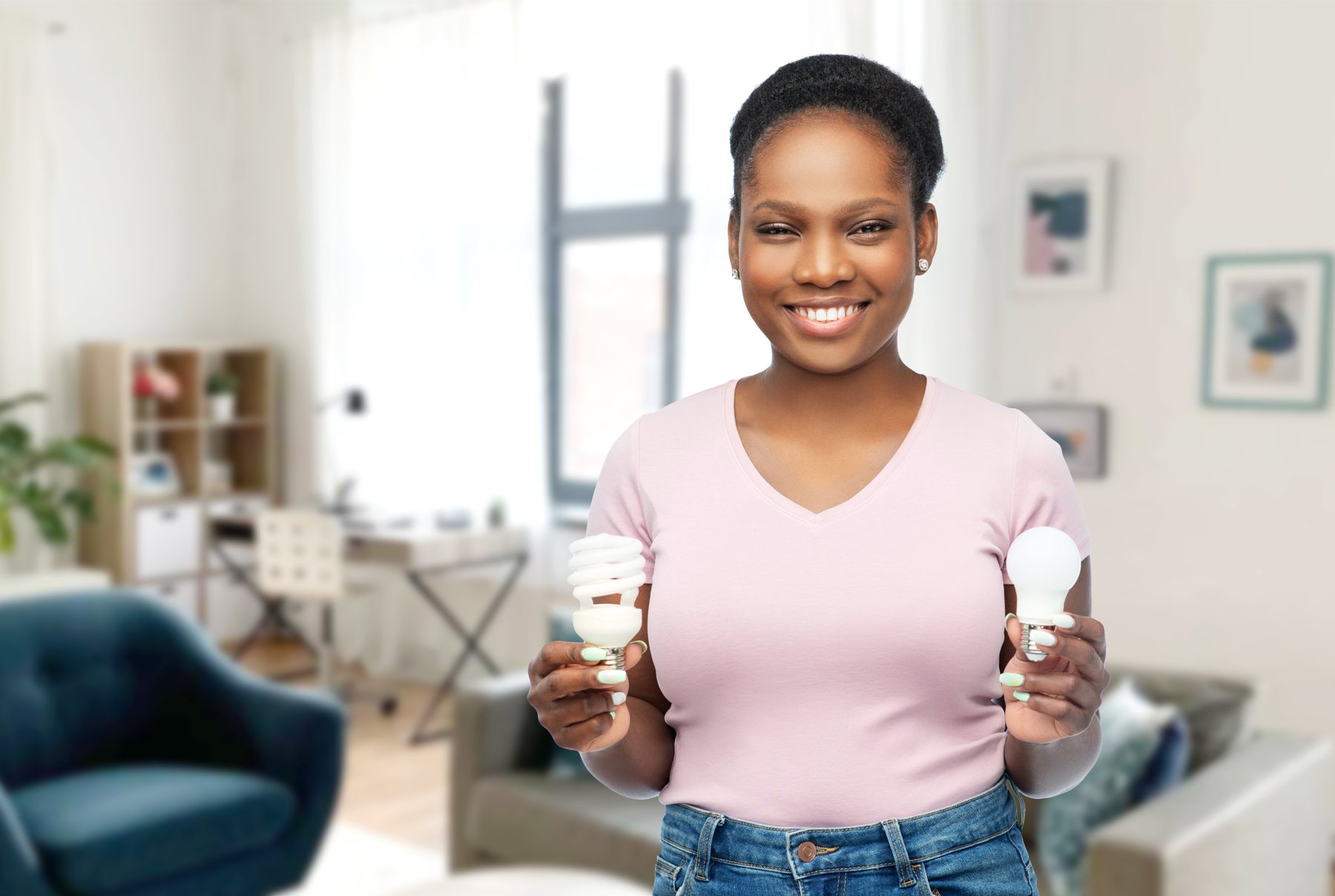 eco living, inspiration and sustainability concept - portrait of happy smiling young african american woman comparing lighting bulbs over home room background