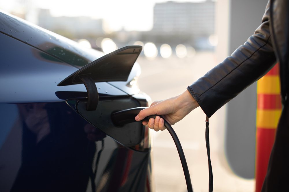 Cropped view of female hand, holding power cable supply plugged in electric car, charging automobile battery from city EV charging station